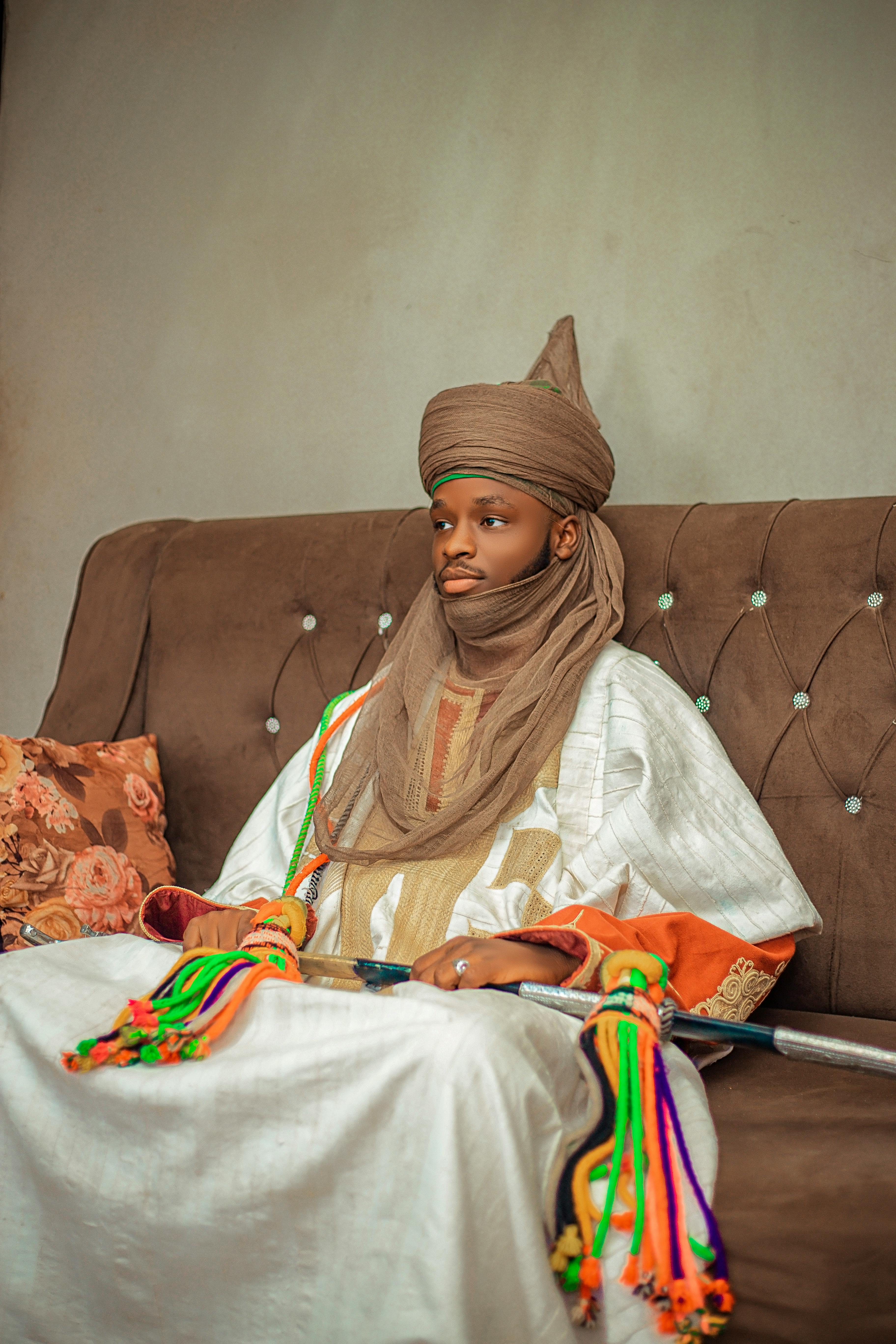 Traditional African emir with elaborate attire seated on a sofa showcasing cultural heritage.