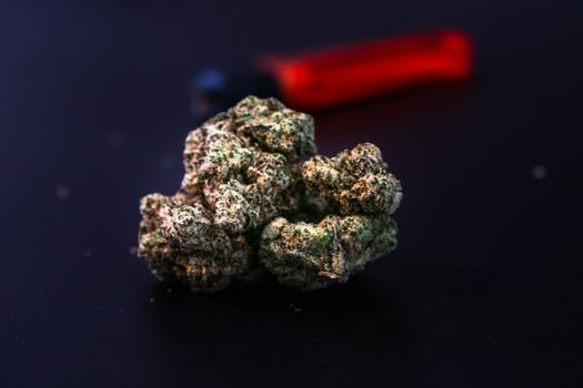 Detailed close-up of a cannabis bud with a red lighter in the background on a dark surface.