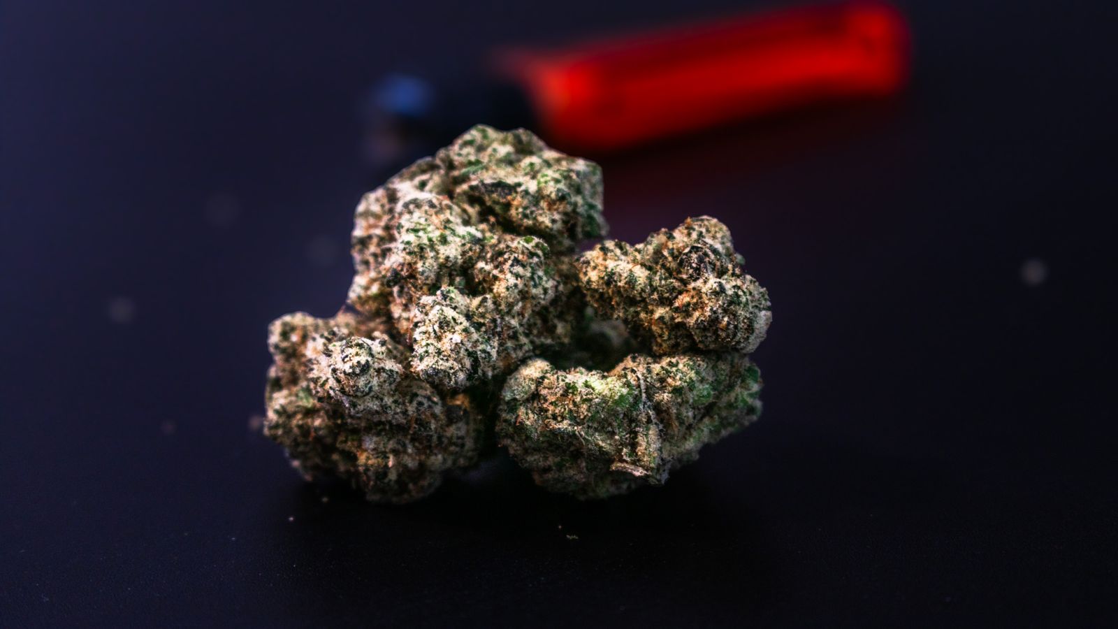 Detailed close-up of a cannabis bud with a red lighter in the background on a dark surface.