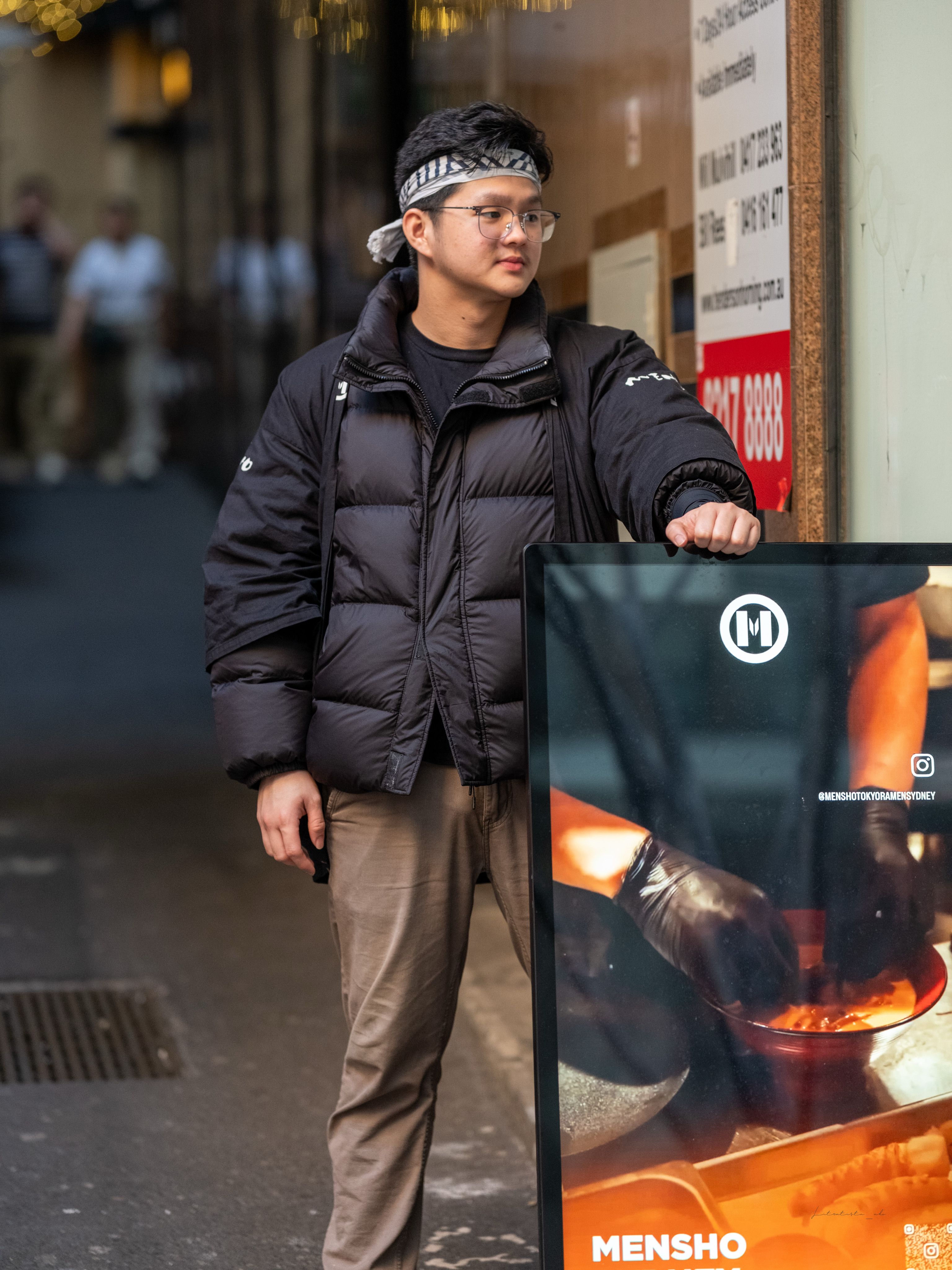 Man in warm clothing stands near restaurant advertisement on busy city street.