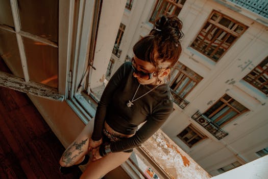 Young woman with tattoos and sunglasses seated on a window ledge in an Argentine building.