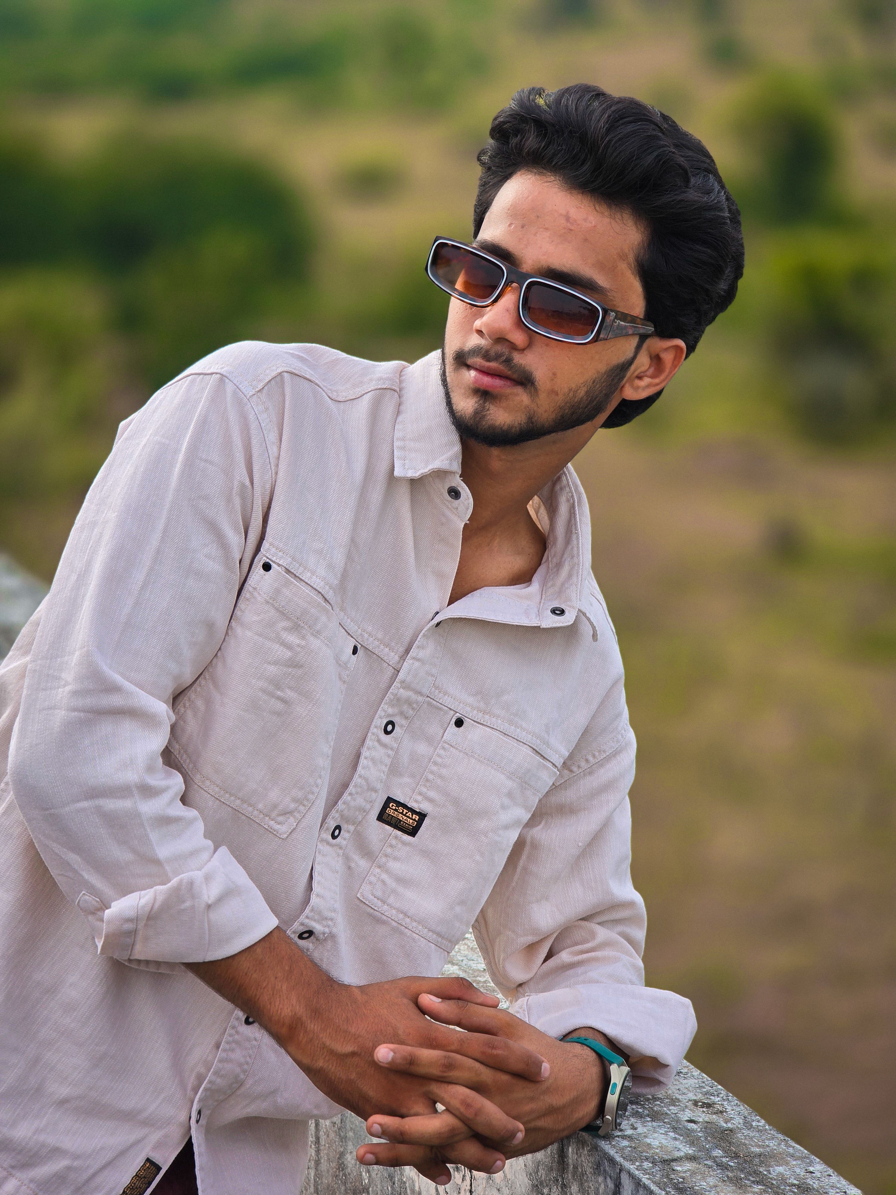 Stylish young man wearing sunglasses and casual shirt, posing outdoors against a natural backdrop.