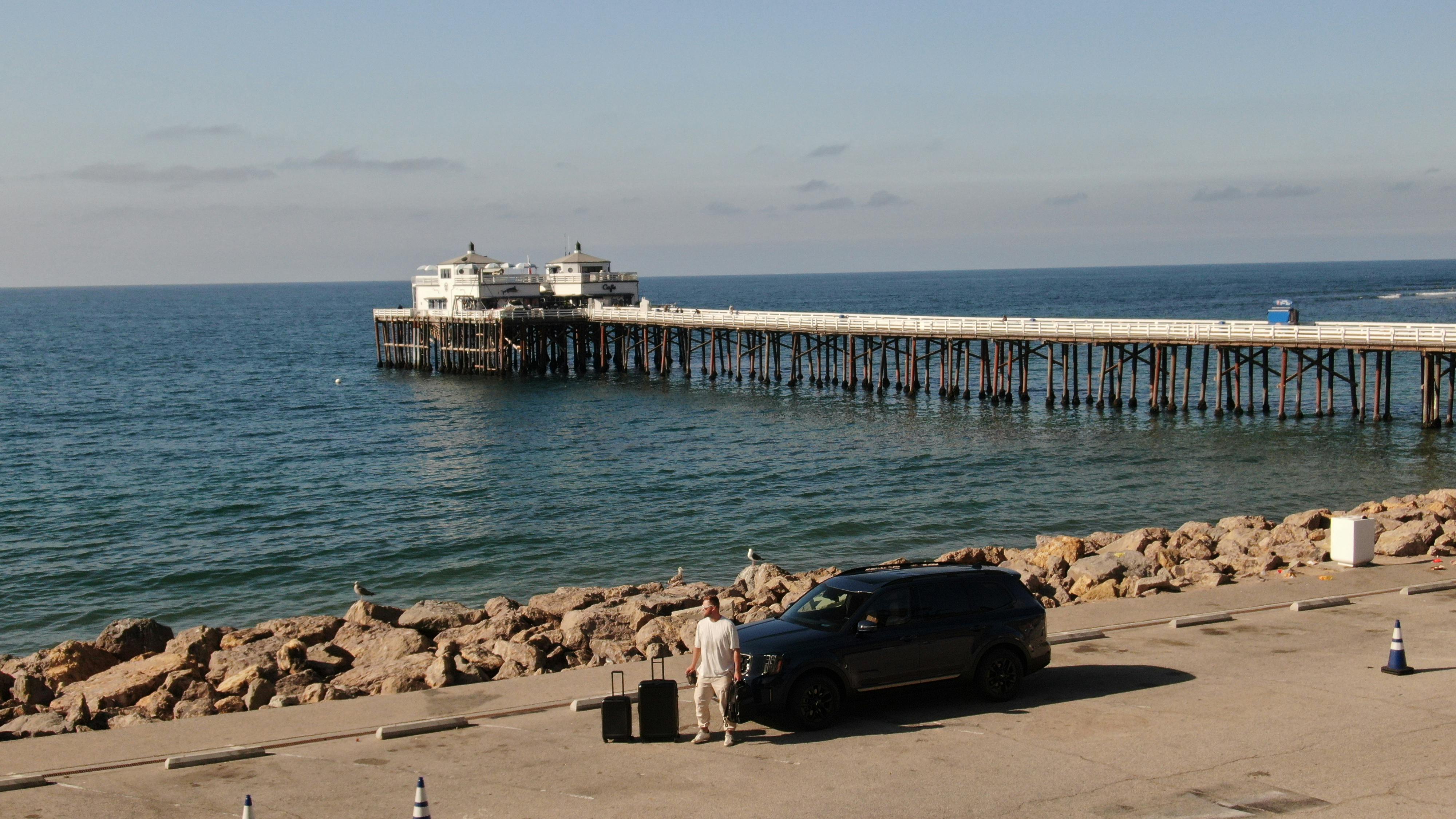 A sunny day at Malibu Pier showing a man with luggage next to a vehicle, overlooking the ocean.