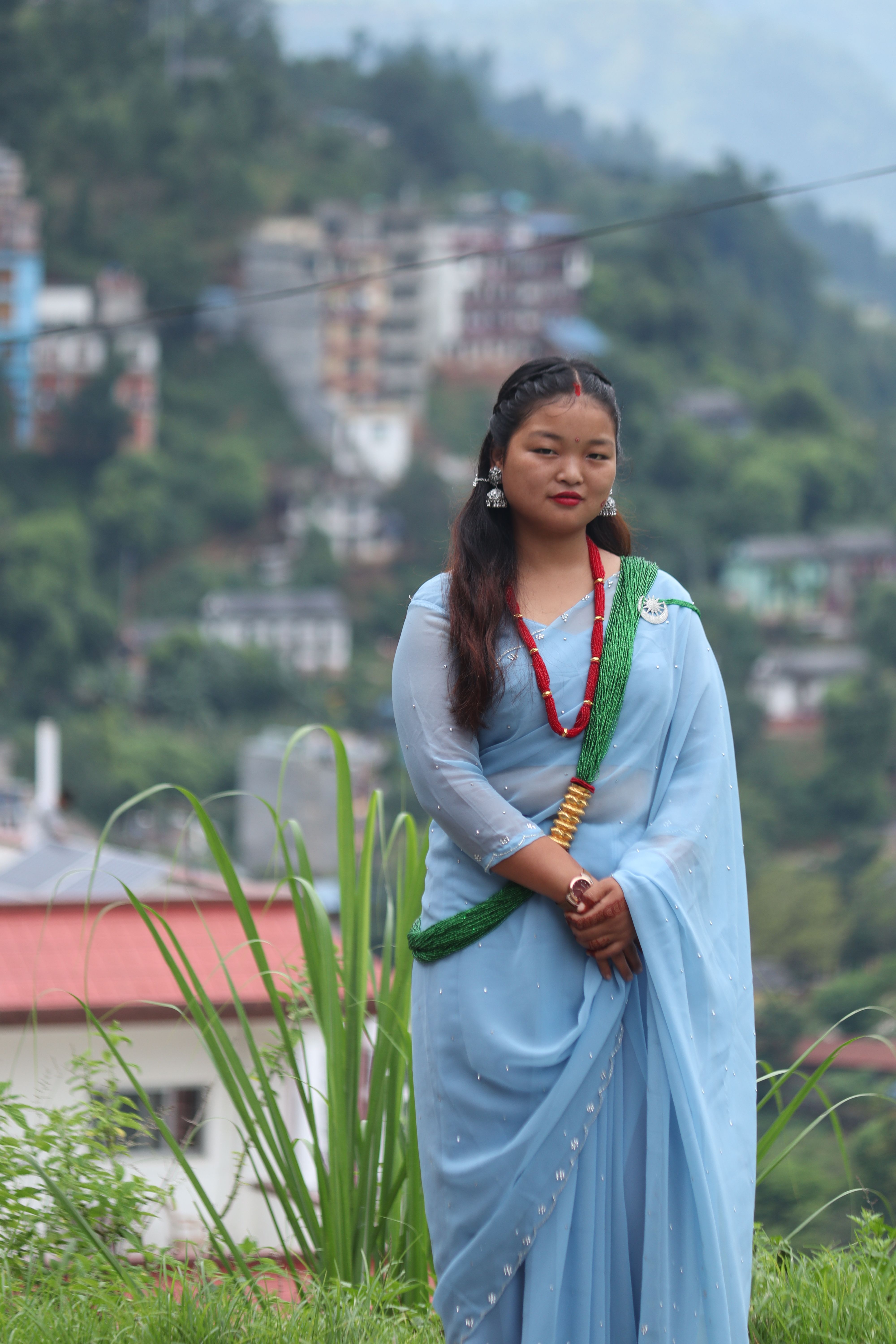 Portrait of a young woman in traditional attire standing outdoors with a scenic hill town backdrop.