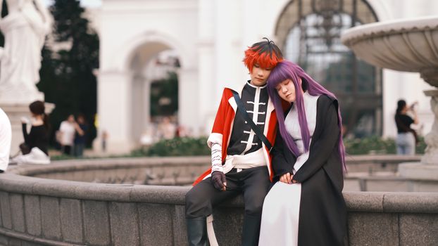 Cosplay couple in vibrant costumes sitting on a stone wall outdoors, embodying anime characters.