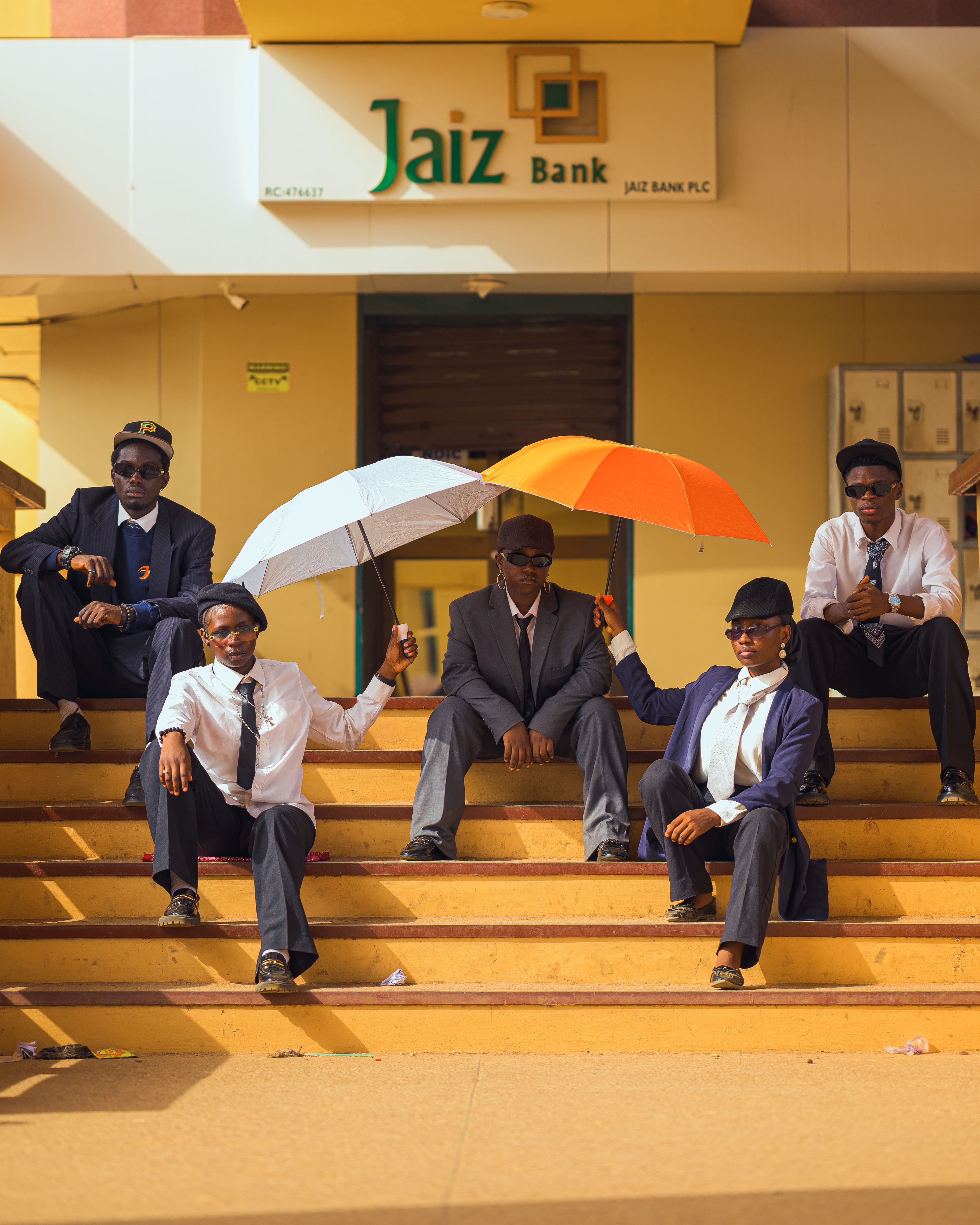 Five young men in suits with umbrellas sitting on steps outside Jaiz Bank, showcasing trendy fashion.