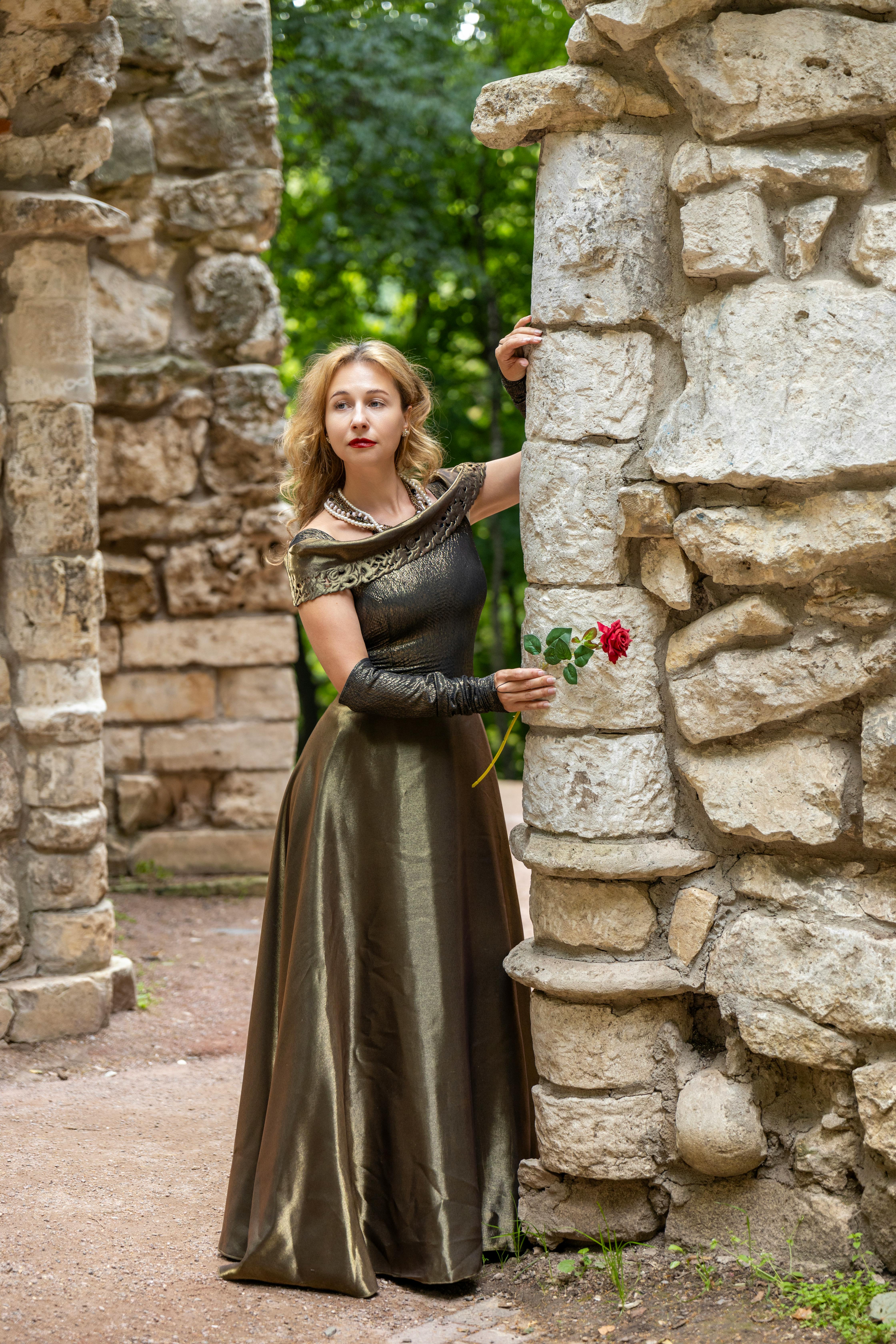 A woman in an elegant gown holds a red rose by ancient stone ruins outdoors.
