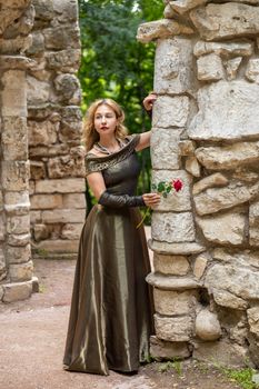 A woman in an elegant gown holds a red rose by ancient stone ruins outdoors.