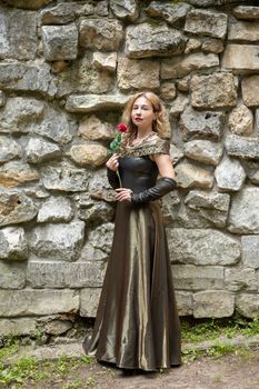 A graceful woman in a vintage gown holding a rose against a rustic stone wall outdoors.