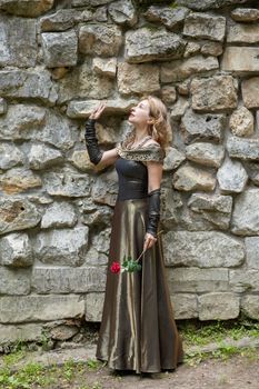 Elegant woman in a medieval-style gown holding a red rose against a stone wall outdoors.