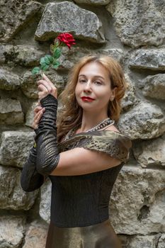 Elegant woman in vintage dress holding a red rose against a stone wall. Medieval style.