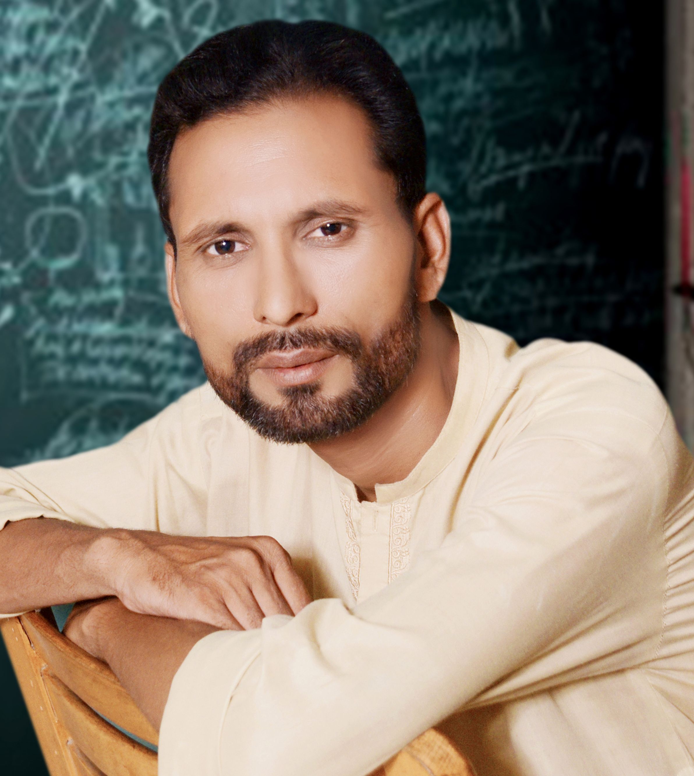 A thoughtful man in a Lahore classroom, against a chalkboard backdrop.