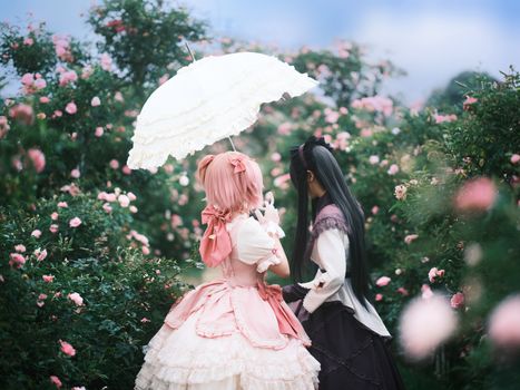Two cosplayers in a garden, dressed in elegant outfits, share a moment under a parasol.