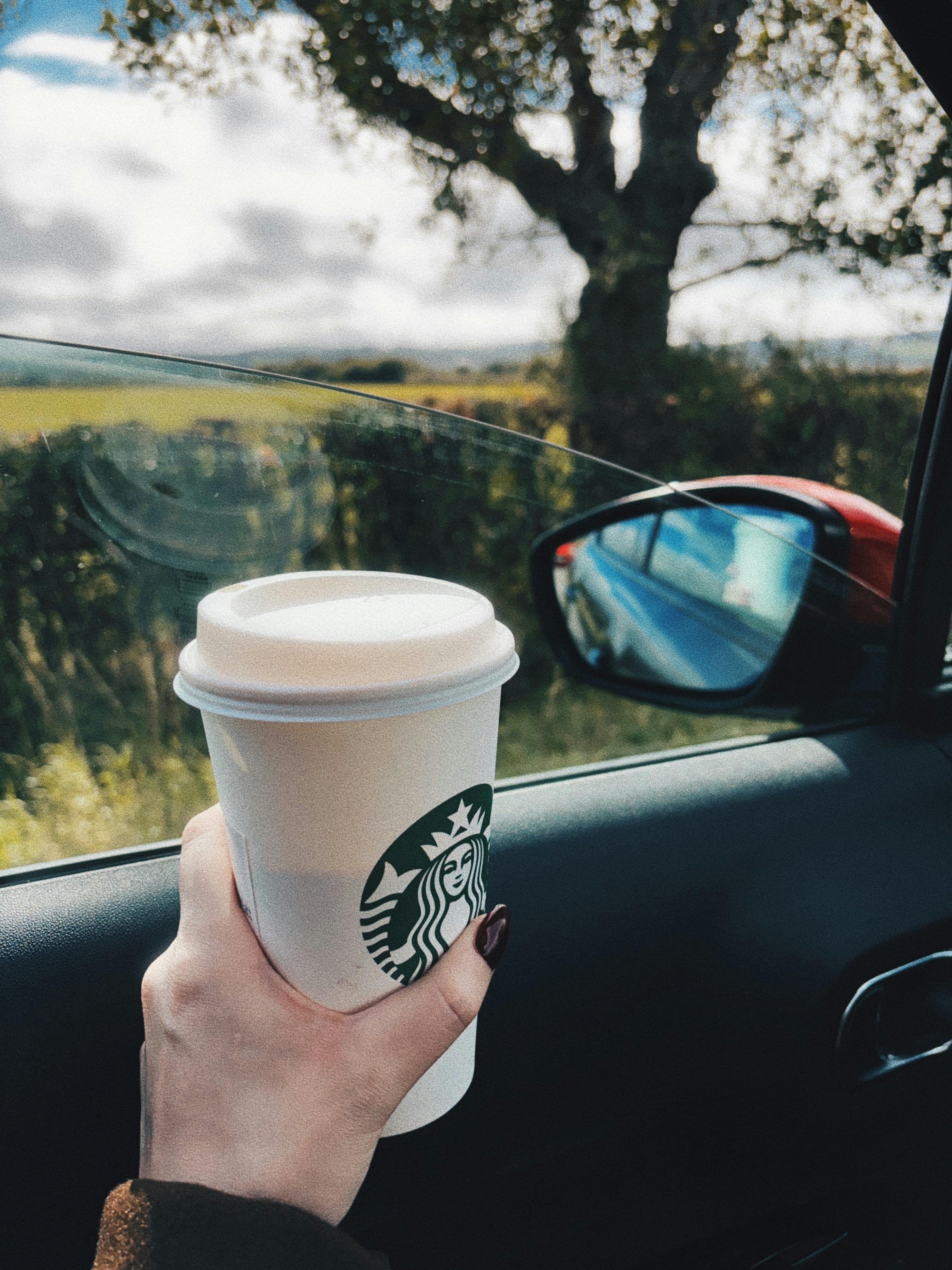 Hand holding coffee cup in car with countryside view outside.