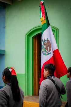 Students in uniform carry the Mexican flag during a National Day parade, with colorful buildings in the background.