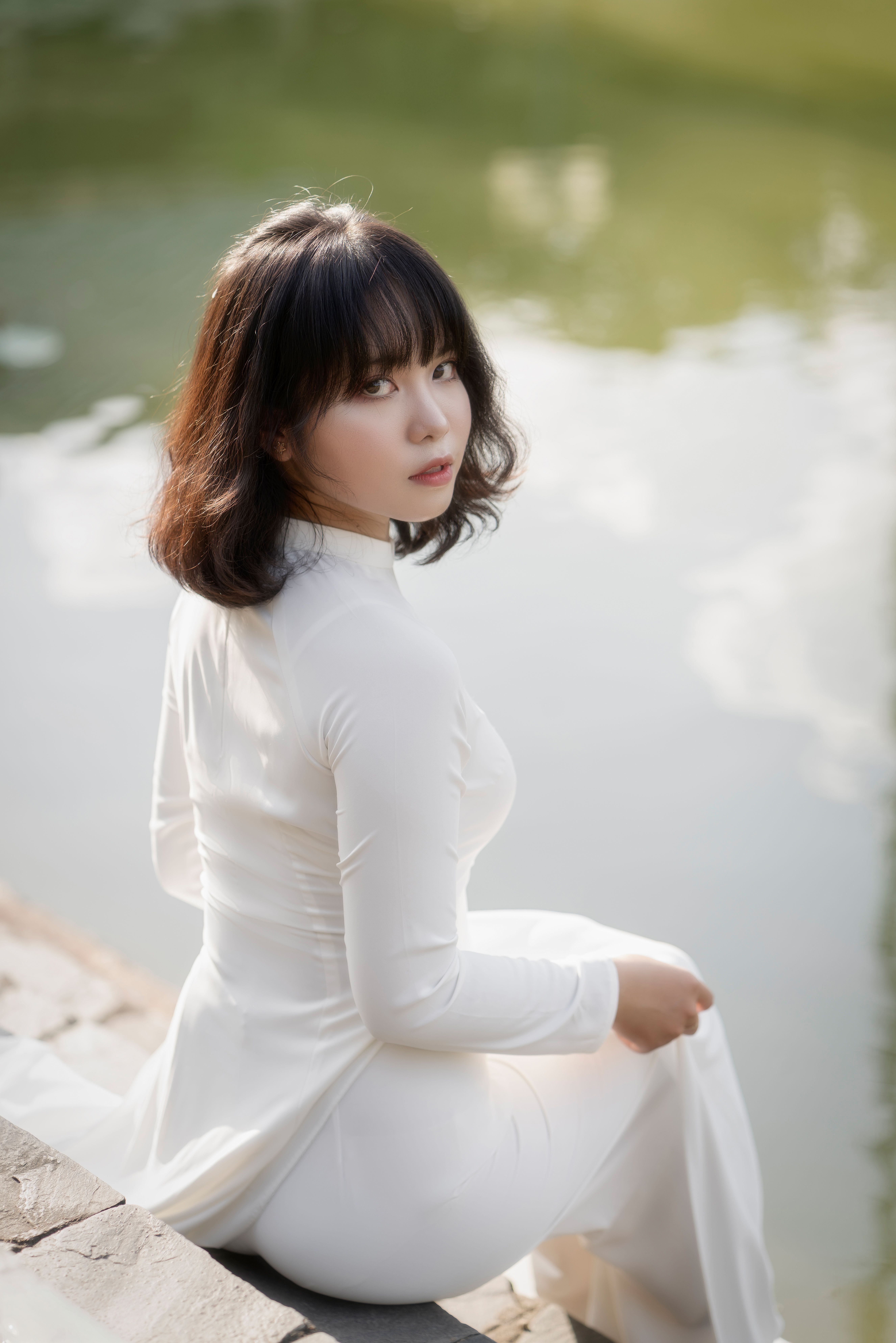 Graceful asian woman in white dress sitting beside serene water reflecting tranquility.