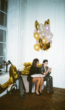 Two women in glamorous attire enjoy a party with festive balloons and stylish decor.