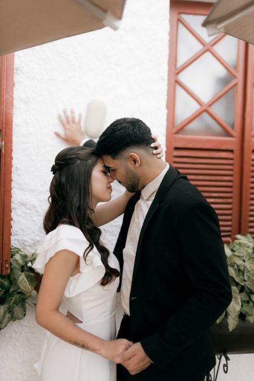 Free Elegant couple in an intimate embrace against a white wall with red shutters. Stock Photo