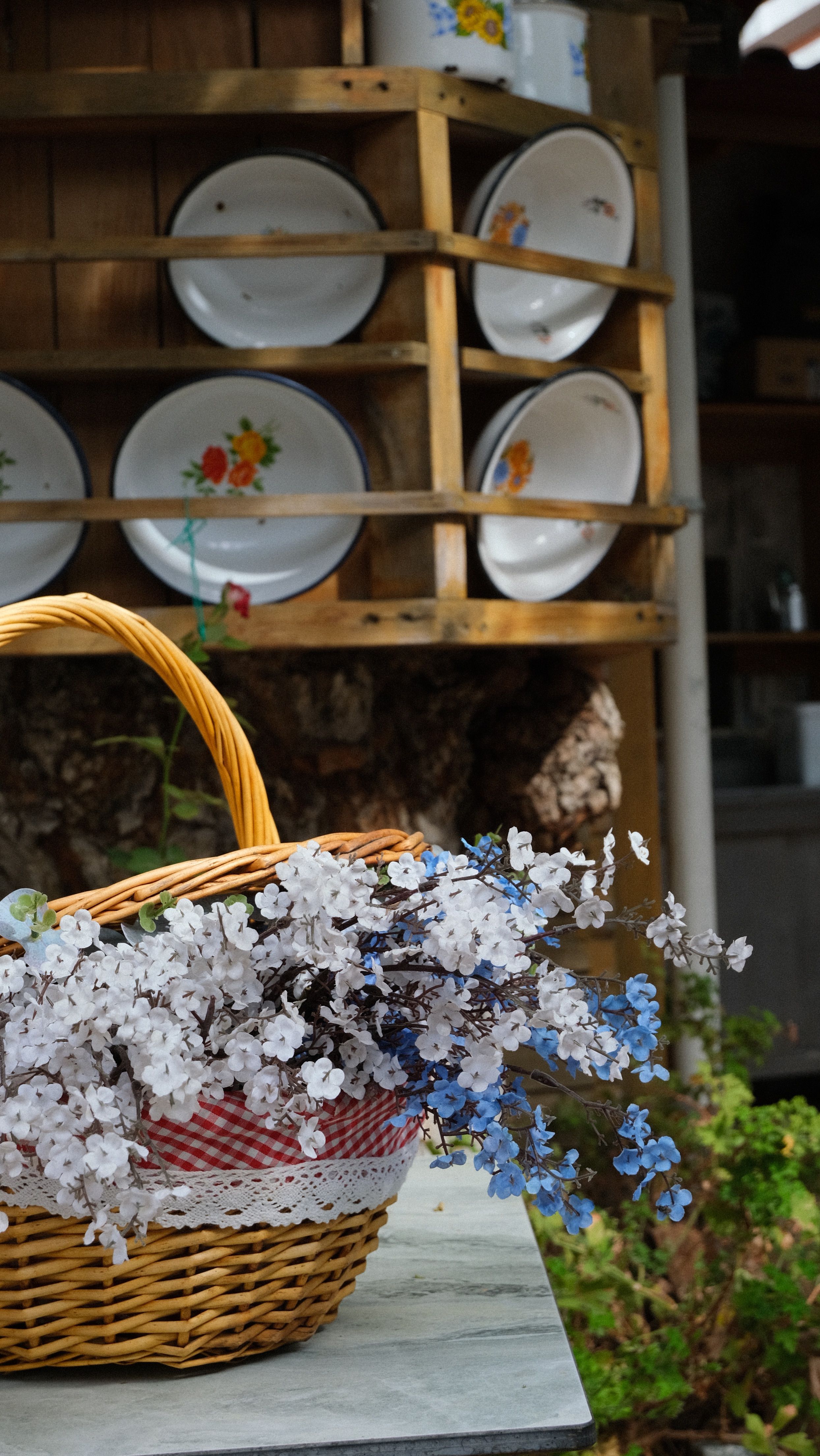Wicker basket of delicate flowers on a table against a vintage plate shelf.