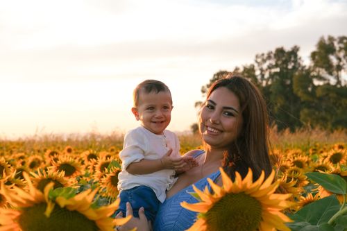 Free A smiling mother holds her baby surrounded by vibrant sunflowers at sunset. Stock Photo