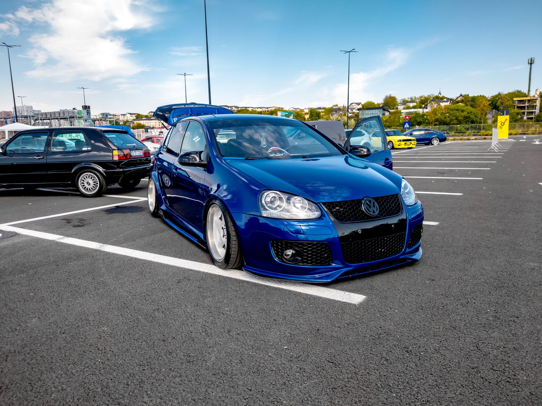 Custom blue Volkswagen Golf parked outdoors with other modified cars in urban environment.