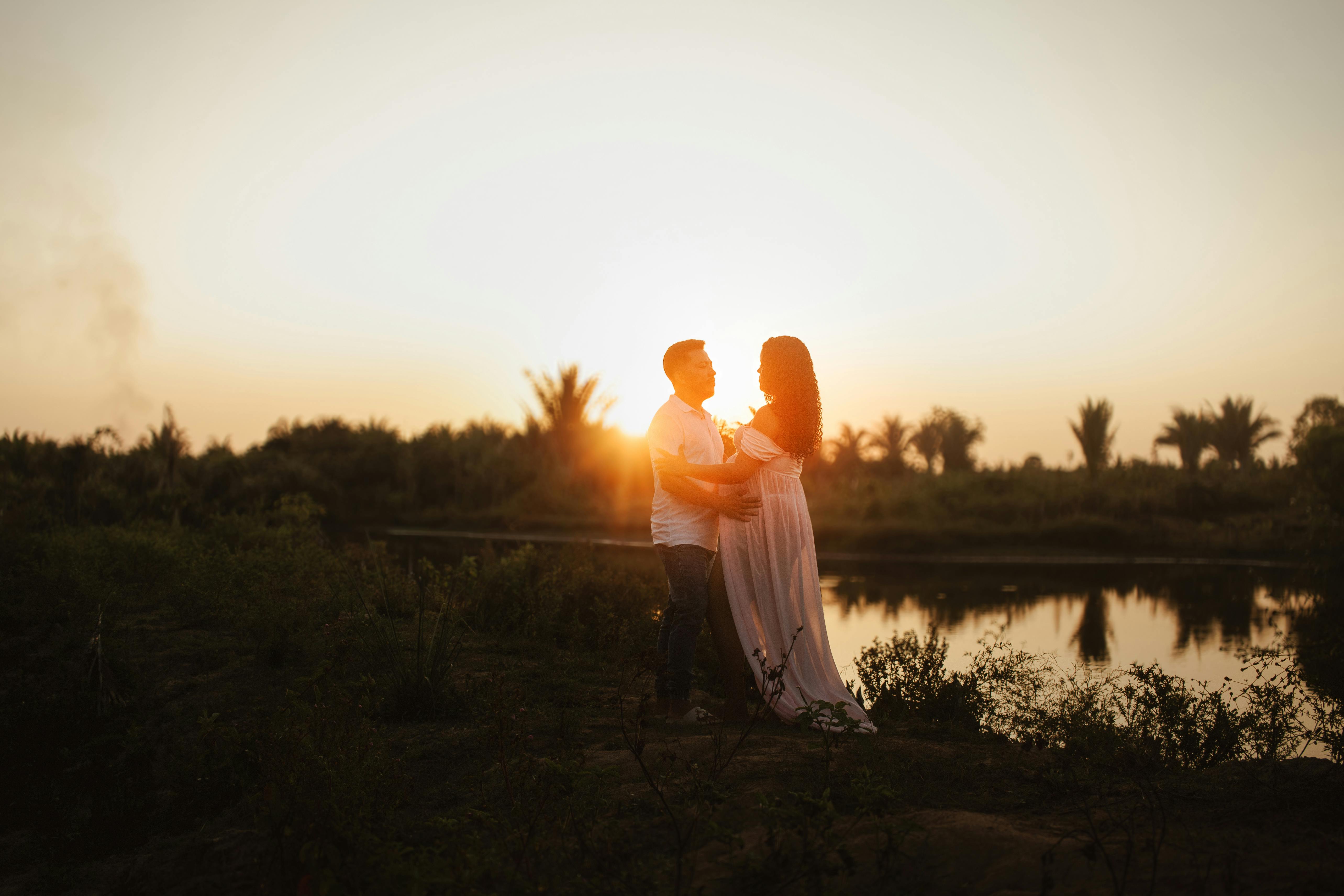Expecting couple embraces by a serene river at sunset, symbolizing love and new beginnings.