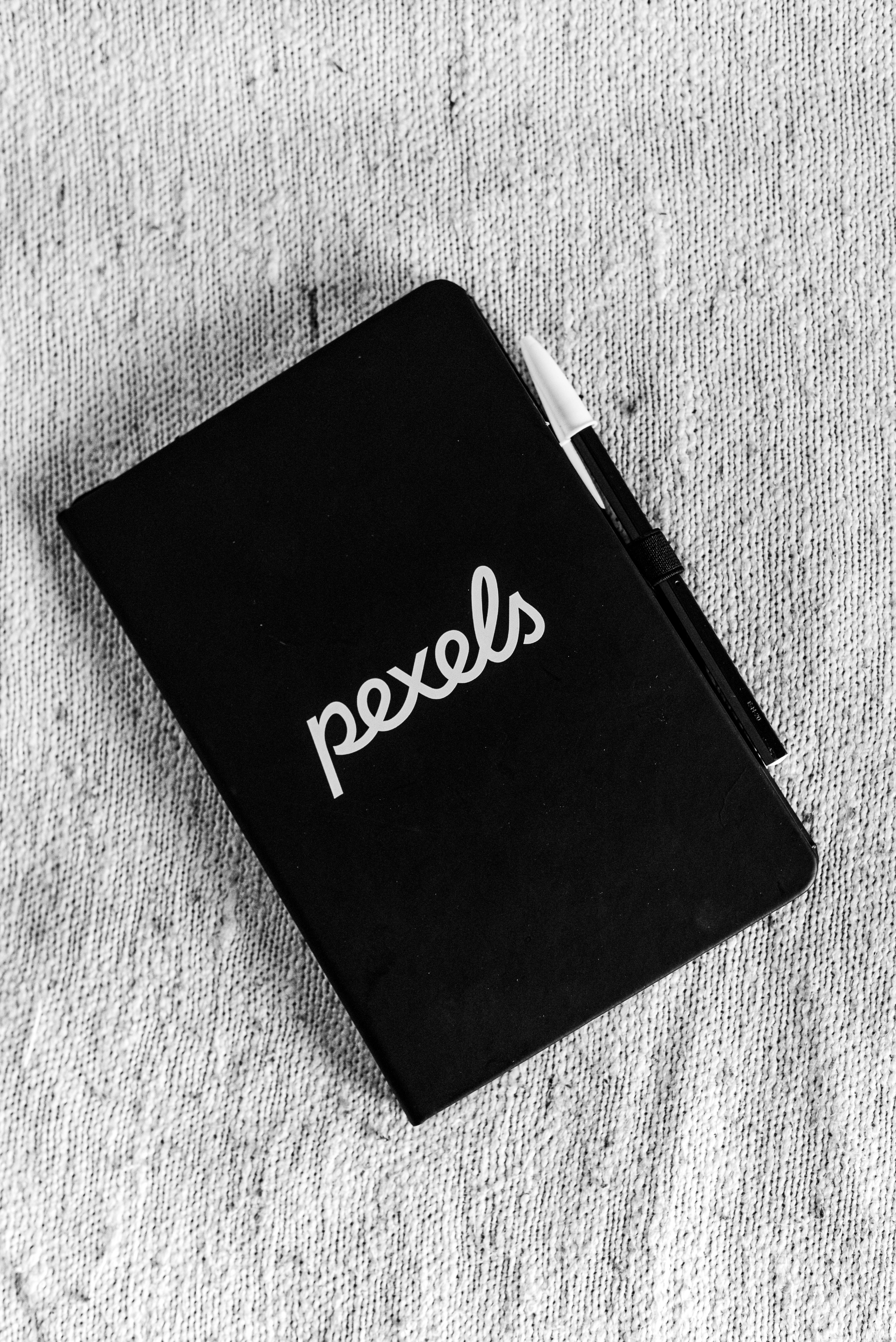 Gratis Monokromt bilde av en lukket svart notatbok med en penn plassert på teksturert stoffbakgrunn. Arkivbilde