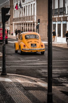 A classic yellow car driving on a city street with historical architecture and pedestrians.