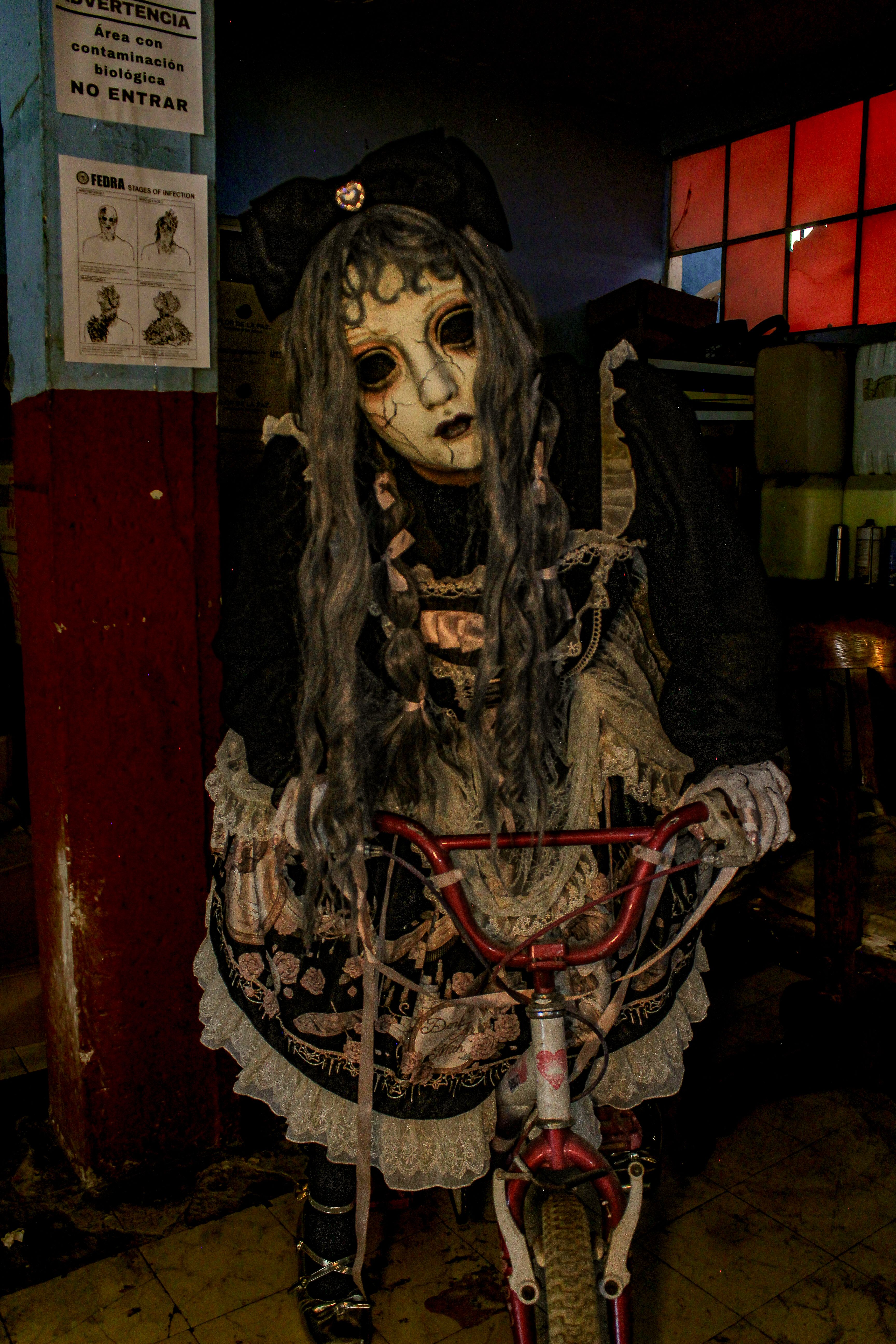 A haunting figure in a detailed mask and costume holding a bicycle in a dim room.