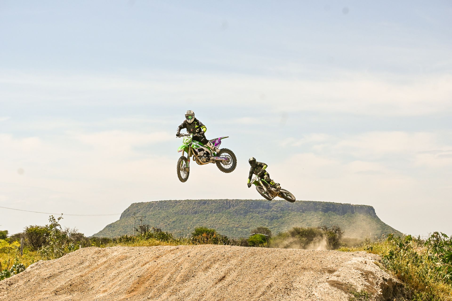Two motocross riders perform daring jumps on dirt bikes against a scenic backdrop.