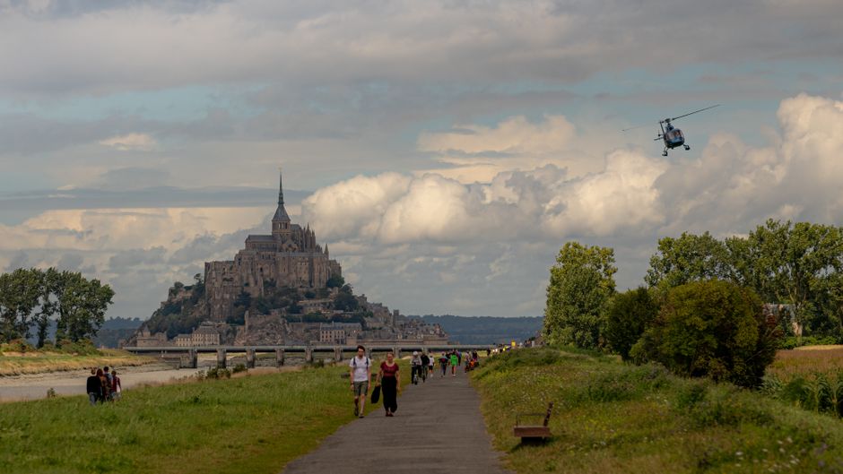 Picturesque landscape with Le Mont-Saint-Michel against dramatic skies, featuring visitors and a helicopter flyby.