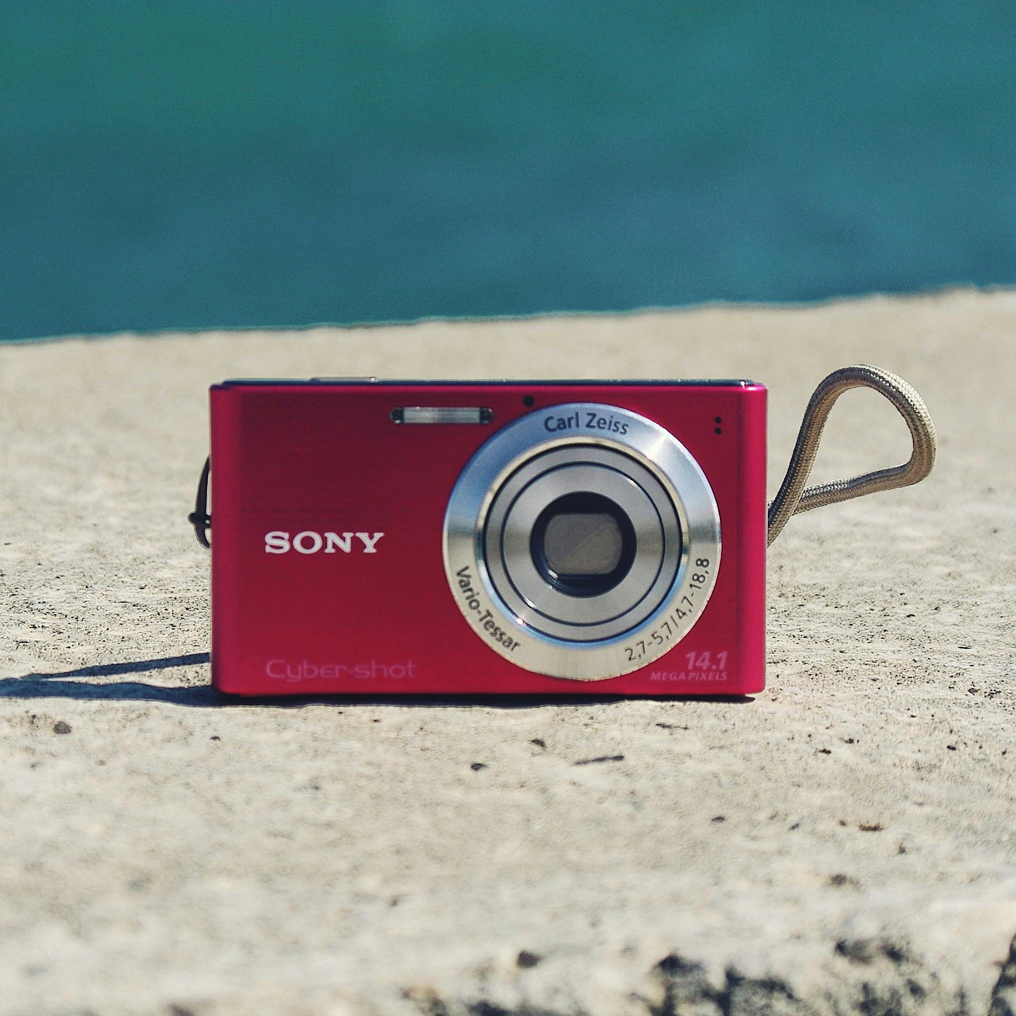 Close-up of a red digital compact camera on a concrete surface with blurred background.
