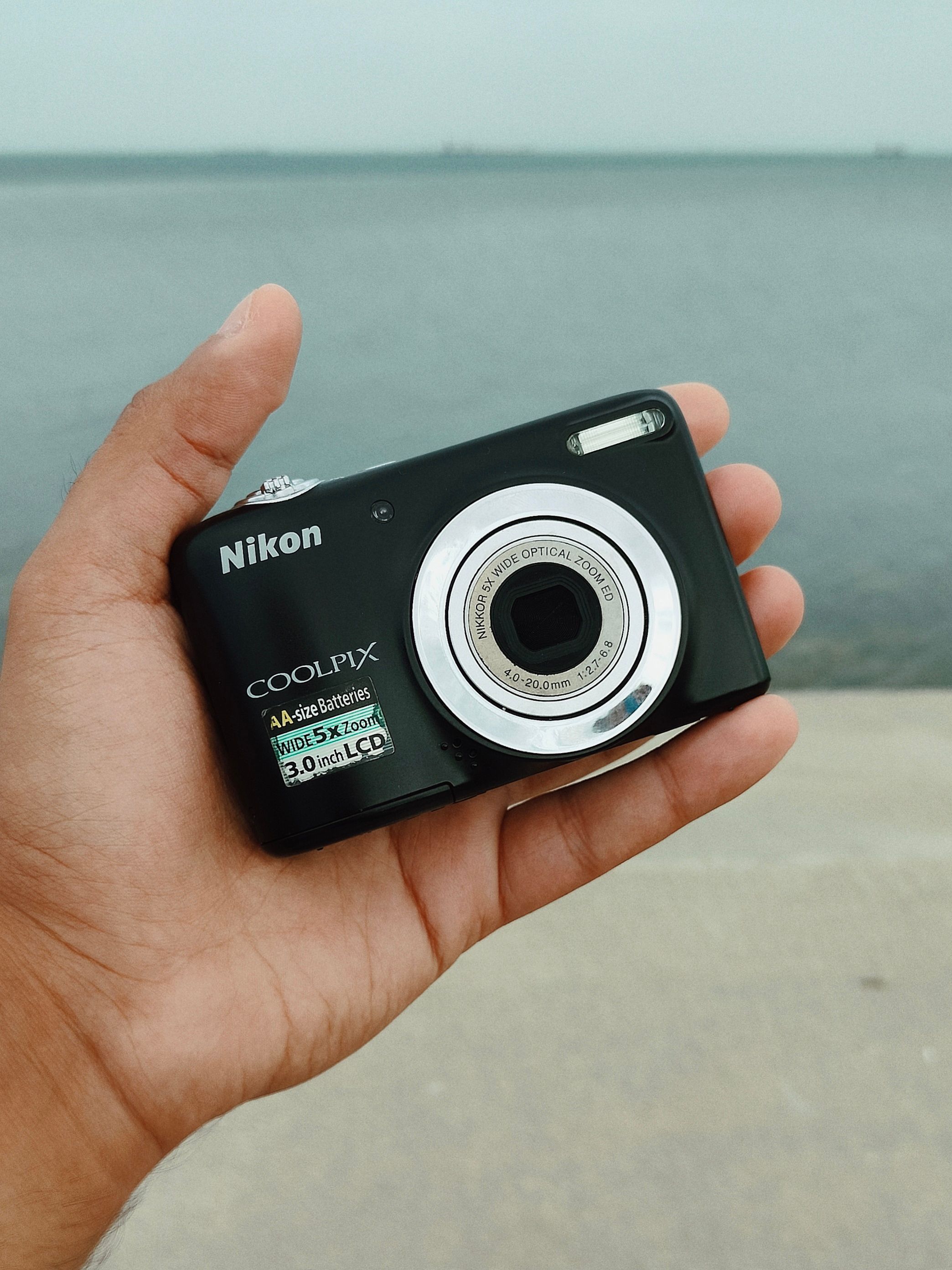 A person holds a black Nikon Coolpix camera outdoors with a serene water backdrop.