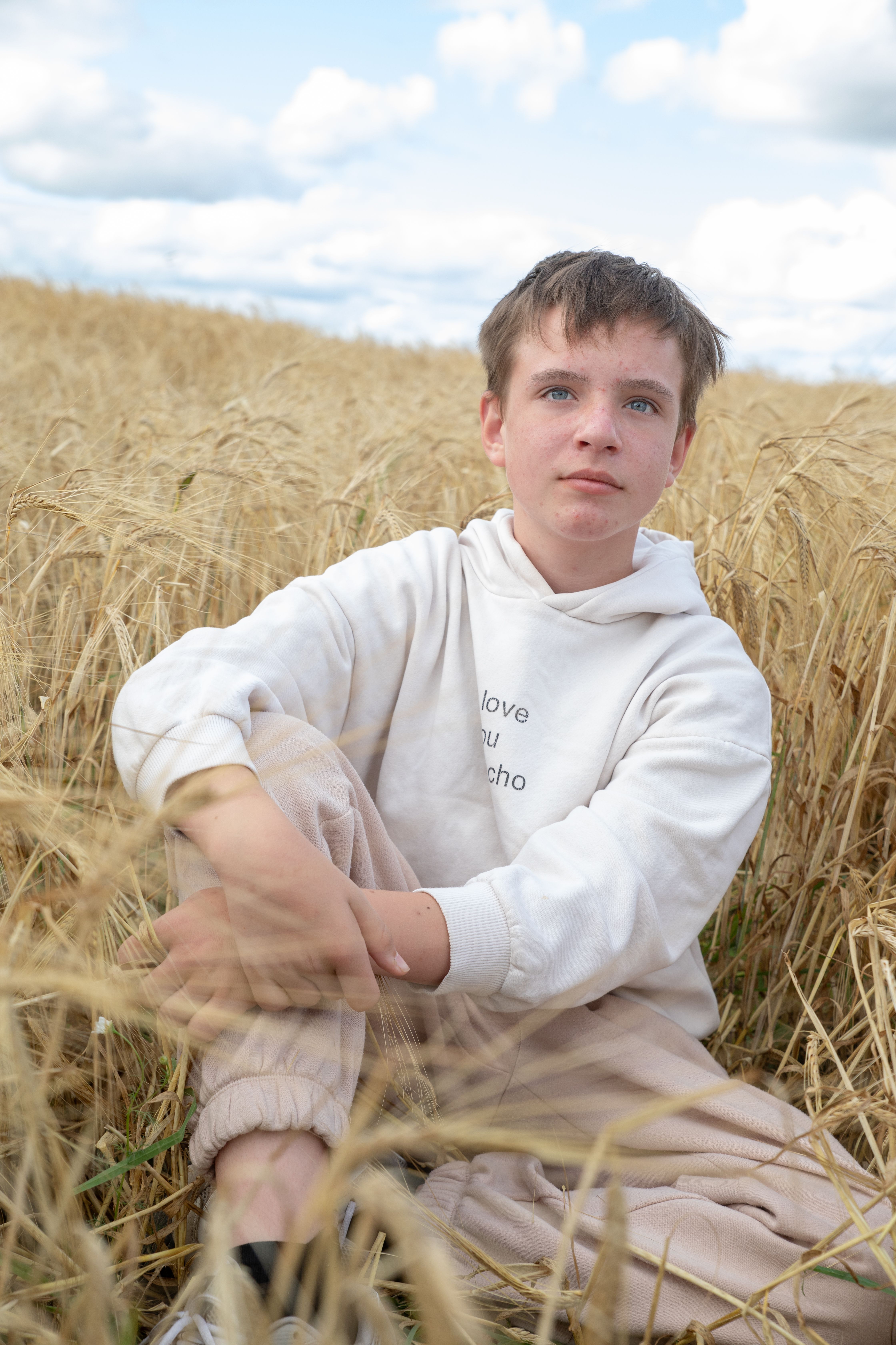 Kostenlos Teenager sitzt nachdenklich in einem goldenen Weizenfeld unter einem bewölkten blauen Himmel. Stock-Foto