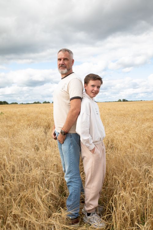 Free Father and son standing back-to-back in a golden wheat field under a cloudy sky. Stock Photo