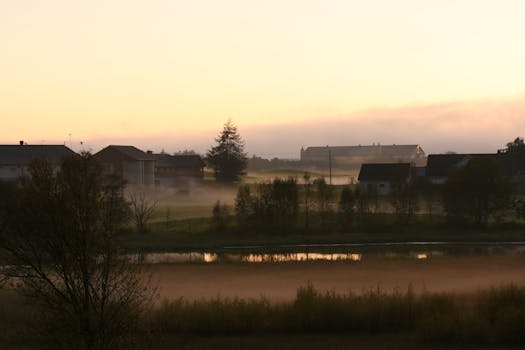 A serene rural scene at sunrise with mist over fields and reflections in a tranquil pond.