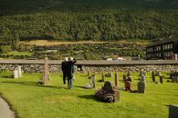 Two men in a peaceful cemetery set against a lush landscape backdrop.