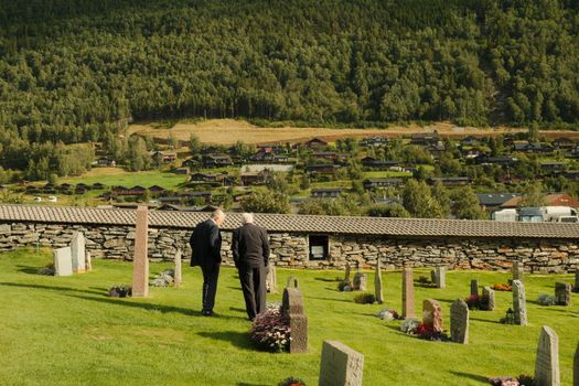 Two men in a peaceful cemetery surrounded by lush hills and rustic homes, capturing a moment of reflection.