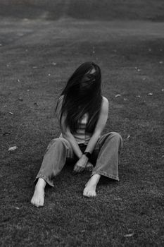A woman sitting alone on grass, her hair covering her face, in a moody black and white setting.