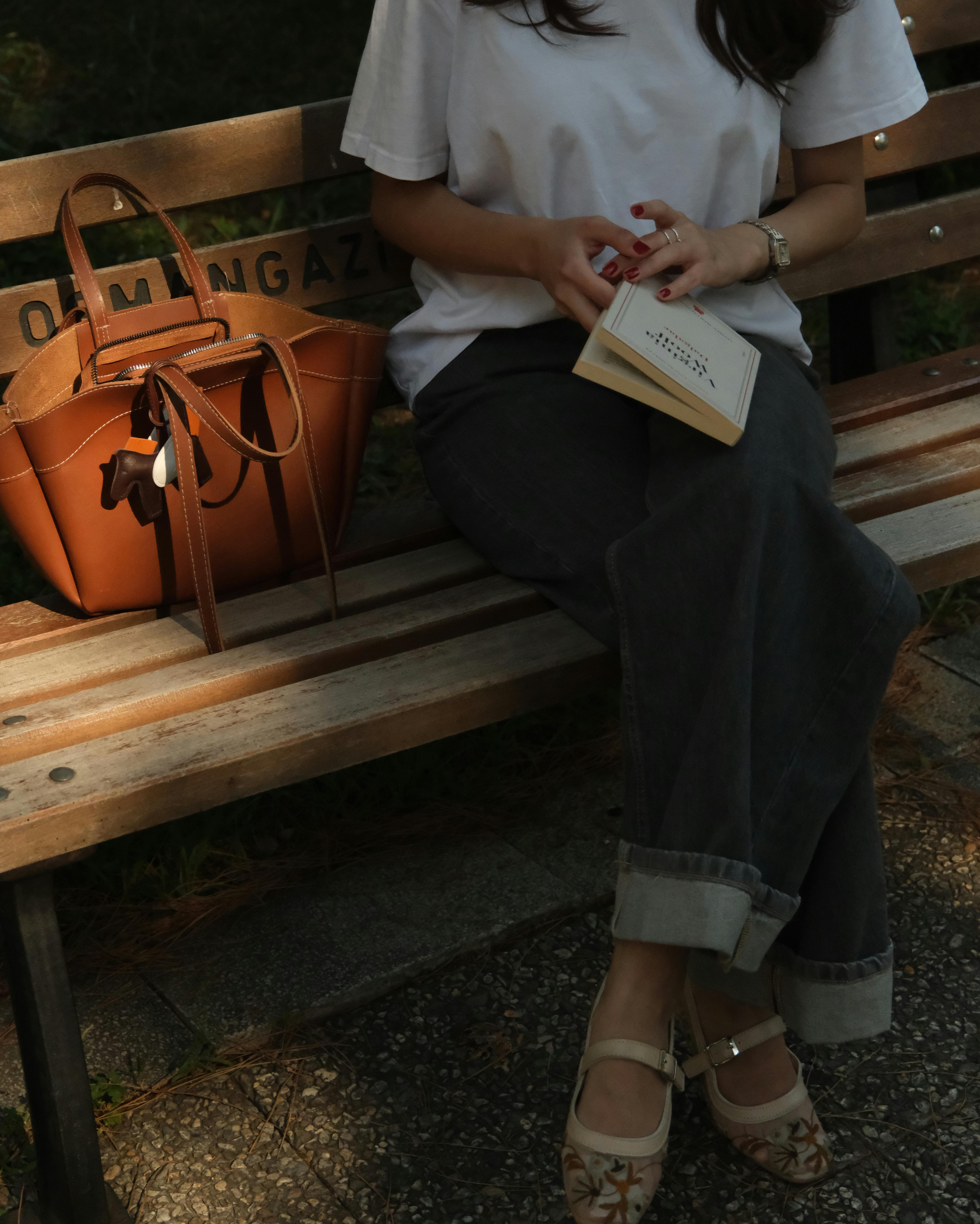 A woman sits on a park bench with a book and brown leather bag, enjoying a peaceful afternoon.