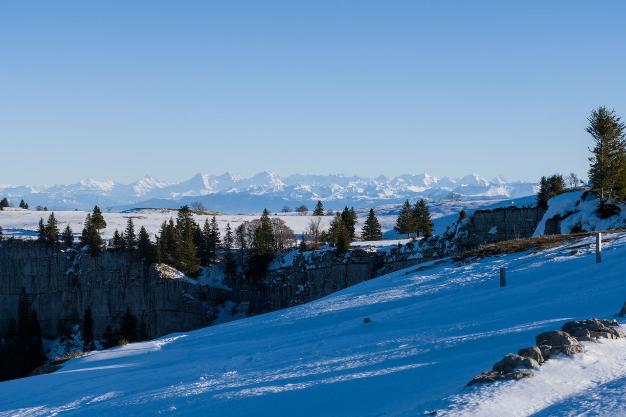 Breathtaking landscape of Creux du Van with snow-covered cliffs and distant mountains under a clear blue sky.