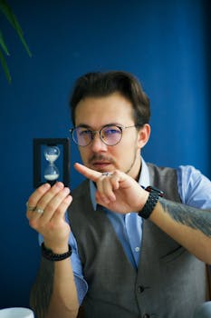 A contemplative adult man holds an hourglass, symbolizing the passage of time, in an indoor setting.