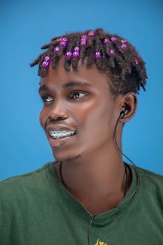 Portrait of a young man with braces, beaded hair, and earphones, set against a blue background.