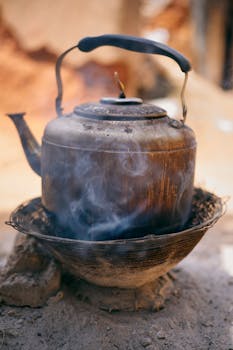 A vintage kettle emits steam as it boils on a rustic outdoor fire pit.