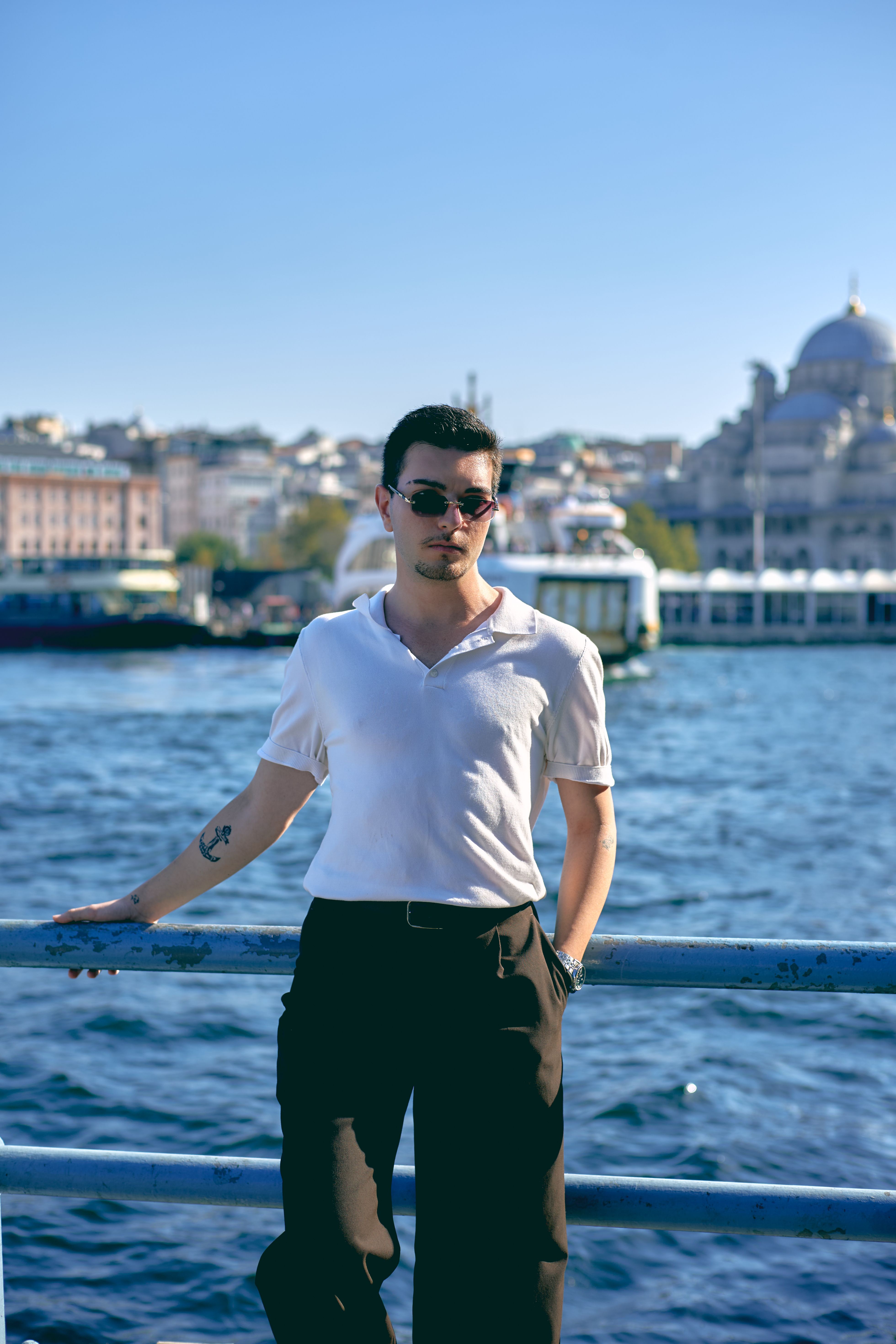 A stylish man poses by the water in İstanbul, featuring the iconic skyline and domes in the background.