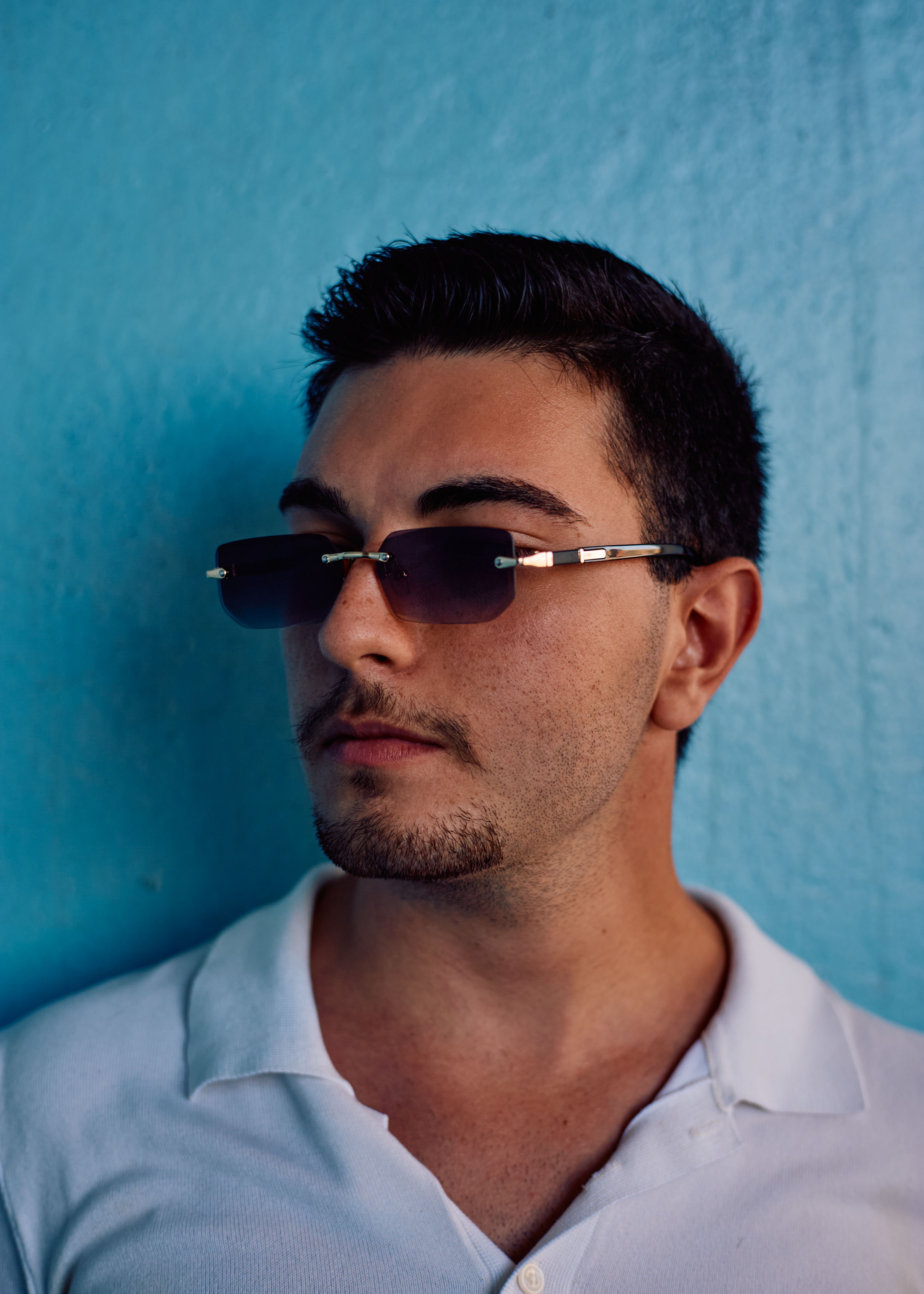 Fashionable adult man wearing sunglasses and white shirt poses against a vibrant blue background outdoors.