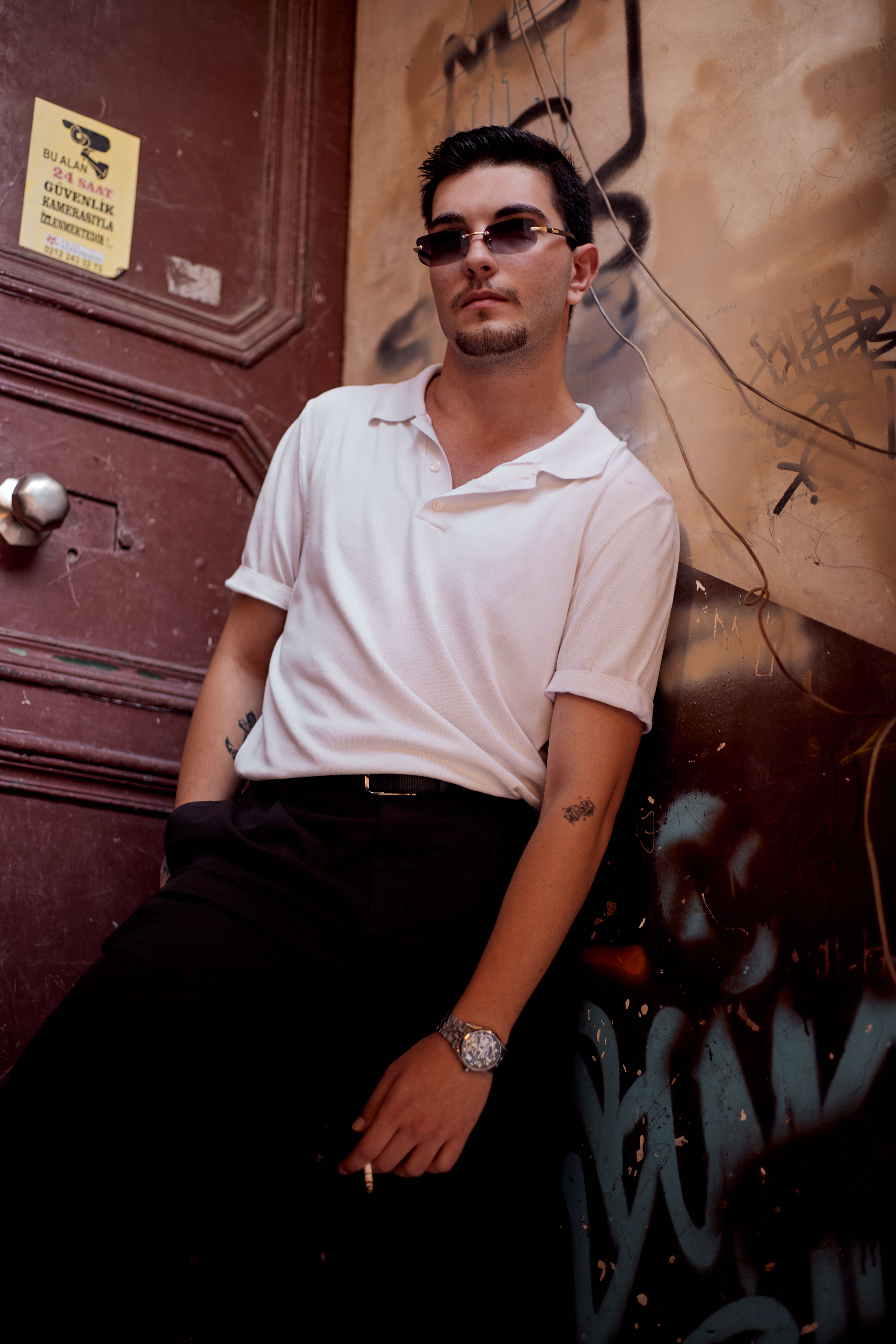 A stylish young man in sunglasses leans confidently against a graffiti-covered wall in İstanbul, Türkiye.