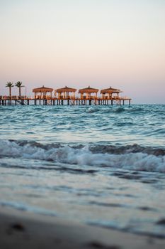 Idyllic beach scene with cabanas on a pier during a tranquil sunset over calm ocean waves.