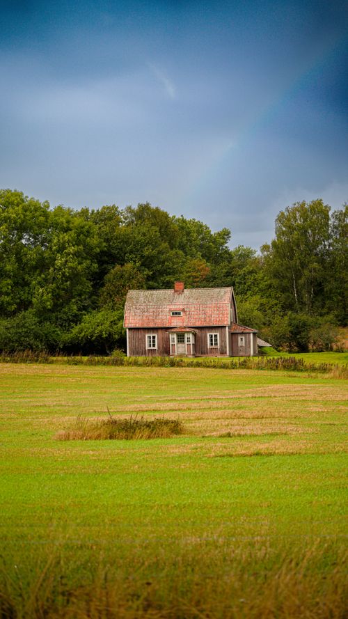 Free Red brick farmhouse amidst verdant field and forest under a dramatic sky. Stock Photo