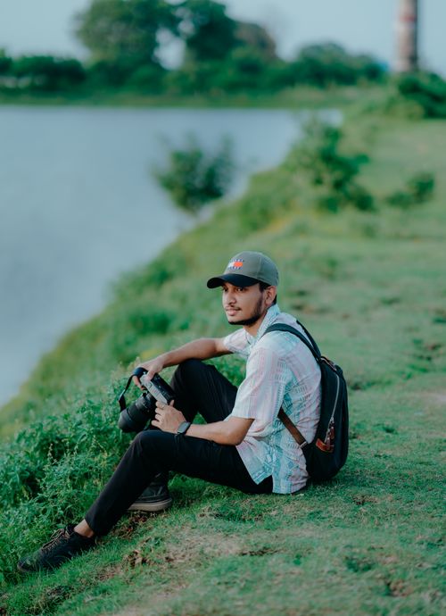 Free A young photographer sitting on a grassy lake shore, enjoying the peaceful outdoor scenery. Stock Photo